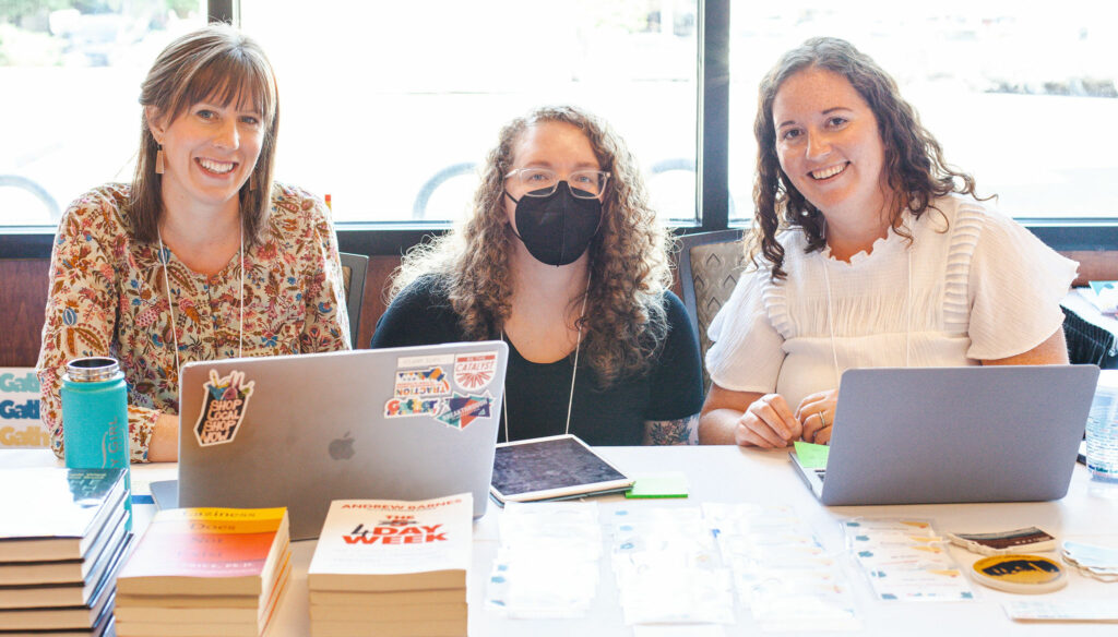 Kate Arpin, Tylyn Newcomb, and Emily Healy pose at a conference registration table, with laptops and stacks of books.