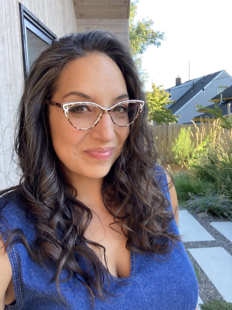 Michelle Muri poses in a backyard, wearing cateye glasses and a blue top. She has long dark brown hair.