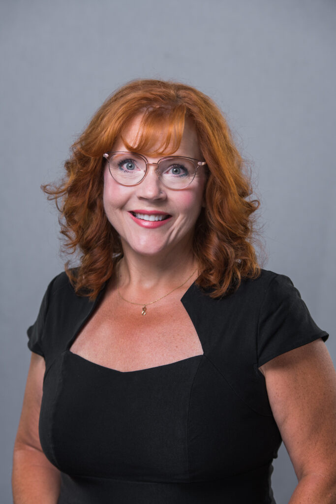 Headshot of Mary Rutherford, a red haired woman wearing glasses and a black shirt.