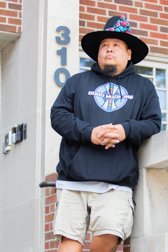 Josiah Hugs leans against a wall, wearing a black hat and a Good Medicine sweatshirt.
